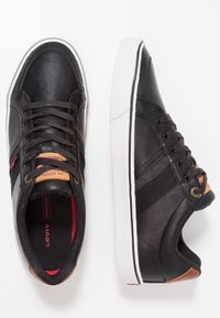 Mustat nahkakengät, joissa valkoiset kumipohjat, ruskeat yksityiskohdat, nauhakiinnitys ja sivulla oleva logotunniste. Pinta on teksturoitu.