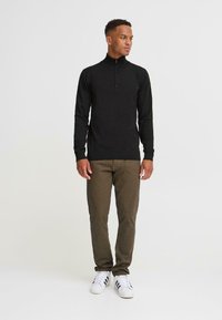 Un jeune homme se tient avec confiance, portant un sweat à zip noir, un pantalon marron et des baskets blanches, sur un fond clair et uni.