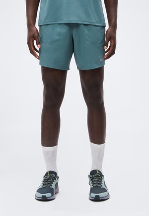 Homme portant un short de sport bleu sarcelle, des chaussettes blanches montantes et des chaussures de course bleu-gris sur un fond uni.