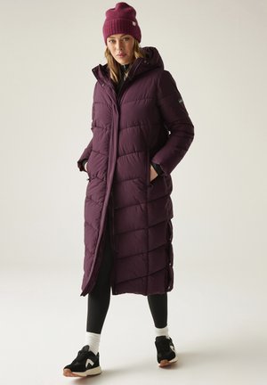 Mujer con un abrigo largo acolchado púrpura con capucha, gorro a juego, mallas negras, calcetines blancos y zapatillas negras, de pie frente a un fondo liso.
