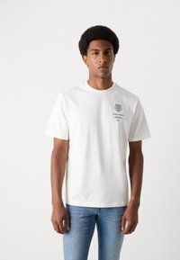 T-shirt bianco in cotone con colletto rotondo. Caratterizzato da una grafica sul petto e maniche corte. Indossato con jeans azzurri chiari. Texture liscia.
