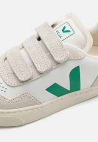 Valkoinen ja vaaleanharmaa lenkkari, jossa on vihreä korostelogo. Siinä on käytetty mokkaa ja nahkaa, Velcro-hihnat sekä teksturoitu kumiulkopohja.