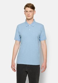 Helles blaues Baumwoll-Polohemd mit einem spitzen Kragen, kurzen Ärmeln und einer kleinen Bruststickerei. Getragen mit schwarzen Jeans.