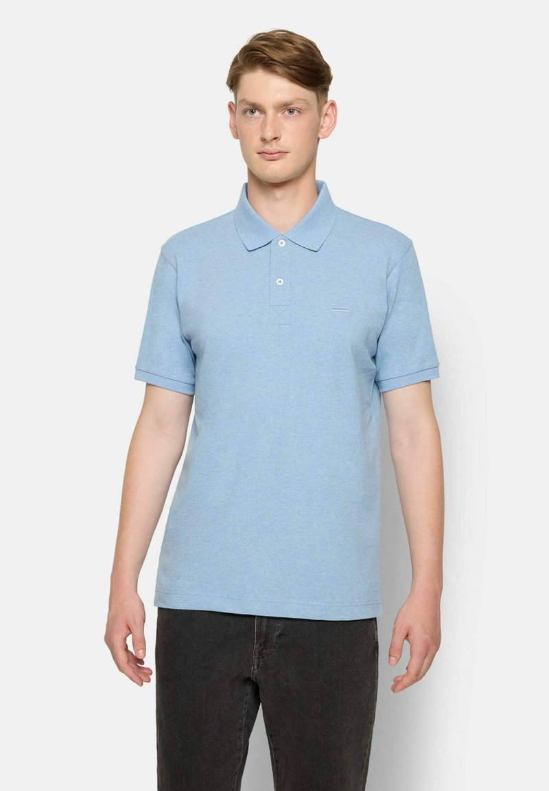 Helles blaues Baumwoll-Polohemd mit einem spitzen Kragen, kurzen Ärmeln und einer kleinen Bruststickerei. Getragen mit schwarzen Jeans.