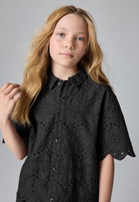 Sort kortærmet skjorte med broderede blomsterudskæringer og hulmønstre, med knaplukning foran og en rynket krave.