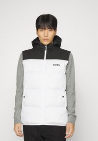 Puikuosių liemenė su balta siuvinėta dalimi, juoda kepure ir pečiais, po kuria matomas pilkas ilgomis rankovėmis marškinėlis su juodais akcentais. Išraiškintas "BOSS" logotipas.