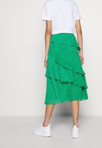 Grön lager midi kjol med volangdesign och blommönster, ihopparad med vita sneakers, sedd bakifrån mot en neutral bakgrund.