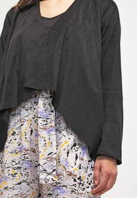 Černý top připomínající semiš s výstřihem do V a asymetrickým lemem, překrytý vzorovanou, barevnou látkou s víry černé, fialové a žluté barvy.