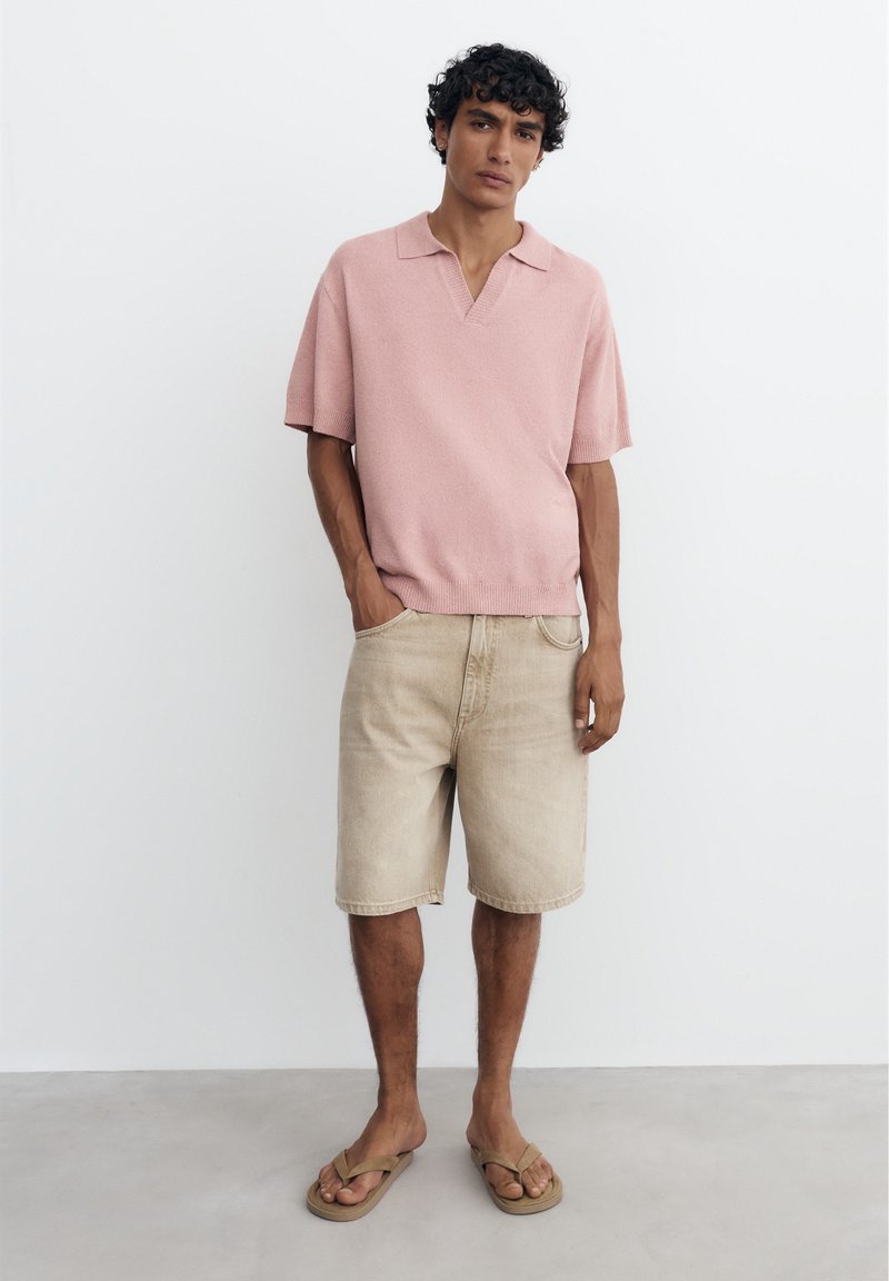 Jeune homme aux cheveux bouclés portant un polo en maille rose à manches courtes, un short en jean beige et des tongs beige, debout contre un mur blanc.