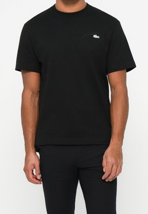 Mężczyzna noszący prostą czarną koszulkę z krótkim rękawem z małym zielonym logo krokodyla Lacoste na lewej piersi.