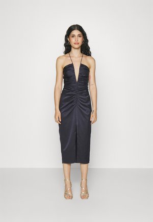 Robe en satin bleu marine avec un décolleté plongeant et des détails plissés. Longueur genou, accentuée par de fines bretelles et une fente sur le devant. Portée avec des escarpins à lanières.