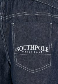 Tissu en denim de couleur bleu foncé avec une poche arrière brodée des mots "SOUTHPOL" et "ORIGINALS" en blanc. Lignes et coutures nettes.