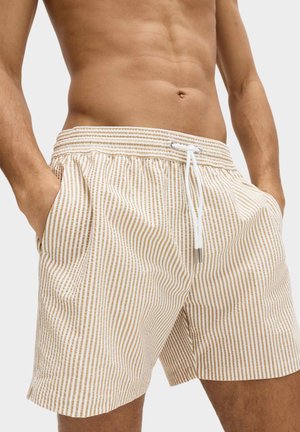 Uomo che indossa pantaloncini da bagno a righe verticali beige e bianche con vita elastica e coulisse bianca, mani nelle tasche.