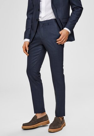 Traje azul marino hecho de un material texturizado, diseño ajustado, con pantalones a juego y una camisa blanca. Mojas marrones con suela gruesa.