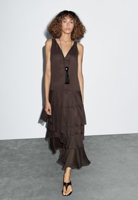 Donna con capelli ricci che indossa un vestito brown senza maniche a strati e una collana con pendente nera, cammina su un pavimento grigio contro un muro bianco.