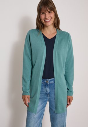 Femme souriante portant un cardigan ouvert couleur sarcelle par-dessus un haut noir et un jean bleu clair, debout devant un fond clair uni.