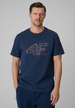 Mężczyzna o krótkich włosach i jasnej brodzie ubrany w granatowy t-shirt z technicznym projektem logo „4F”.
