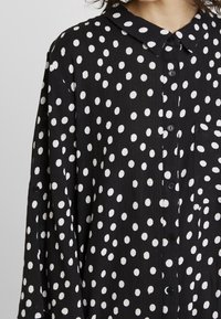Schwarz karierte Hemd mit weißen Polka-Punkten, einem Kragen, langen Ärmeln, einem strukturierten Stoff und einer einzelnen Brusttasche.