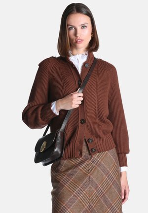 Femme portant un cardigan en maille marron boutonné, une jupe à carreaux, un chemisier blanc à volants, et un sac à épaule en cuir noir sur un fond uni.