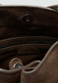 Interior de un bolso de ante marrón con un bolsillo con cremallera, cierre a presión y una etiqueta de marca visible. Textura suave con costuras limpias.
