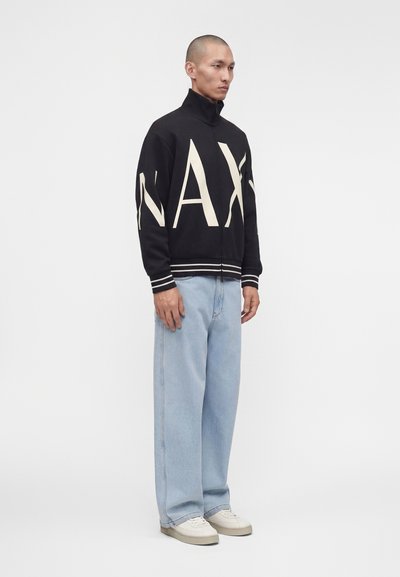 Homme debout portant une veste noire zippée avec de grandes lettres blanches "MAX", jean large bleu clair et baskets blanches, sur fond blanc.