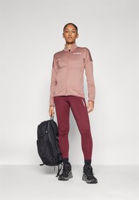 Terrex zip-up jakna u svijetlo ružičastoj boji, bordo tajice i crni ruksak. Teksturirani materijal, prilagođen dizajn i kontrastni logo akcenti.
