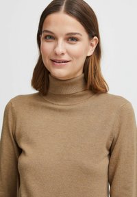 Maglione a collo alto marrone realizzato in morbido tessuto a maglia fine con maniche lunghe e colletto a coste, caratterizzato da una struttura liscia e un design minimale.