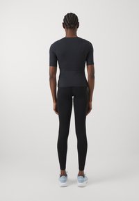 Schwarzes kurzärmliges, figurbetontes Top, das mit schwarzen Leggings kombiniert wird. Das Top hat eine glatte Textur; die Leggings sind dehnbar. Getragen mit hellblauen Sportschuhen.