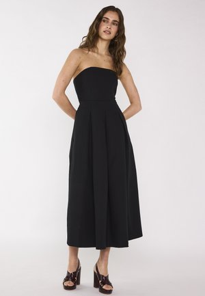 Donna che indossa un abito midi nero senza spalline con pieghe, abbinato a sandali con plateau marroni open-toe, in piedi davanti a uno sfondo bianco semplice.