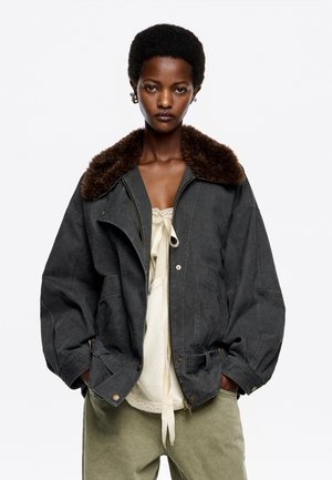 Chaqueta de gran tamaño de color gris oscuro con un cuello de piel marrón, que cuenta con un cierre frontal, bolsillos y detalles en los puños. Lleva puesta sobre una blusa de color beige claro.