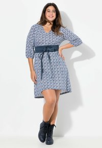 Ein blaues, gemustertes Kleid mit V-Ausschnitt, 3/4-Ärmeln und einem dunkelblauen Gürtel. Kombiniert mit schwarzen Schnürstiefeletten.