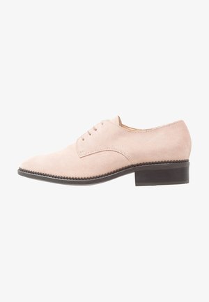Zapato oxford de ante beige claro con cordones cerrados, detalles de costura y un tacón bajo apilado de color marrón oscuro.