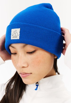 Blue knitted beanie with ribbed texture, featuring a white label with a mountain logo. Worn, partially covering brown hair.