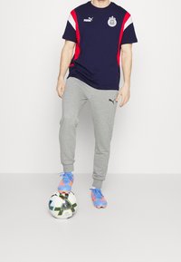 Navy and red short-sleeve sports t-shirt with Puma logo, paired with light gray sweatpants and blue-orange soccer cleats, standing over a soccer ball.