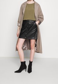Cappotto di lana beige, top verde, gonna di pelle nera con bottoni frontali e stivaletti neri con punta affusolata e tacco alto.