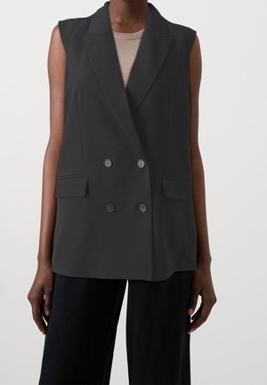 Personne portant un blazer noir sans manches à double boutonnage avec poches à rabat sur un top beige et un pantalon noir sur un fond uni.