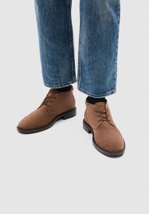 Persona che indossa jeans blu e stivali stringati in camoscio marrone con suole nere, in piedi su uno sfondo grigio chiaro.