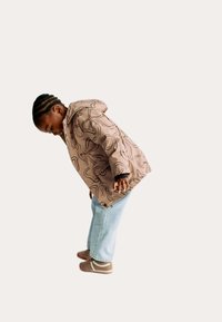 Beige regnjakke med sort abstrakt mønster, udstyret med hætte og lynlås. Kombineret med lyseblå jeans og brune sko.