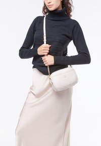 Borsa a tracolla in pelle testurizzata grigia chiara con hardware dorato e doppie zip, indossata con un dolcevita a coste blu navy e una gonna crema.