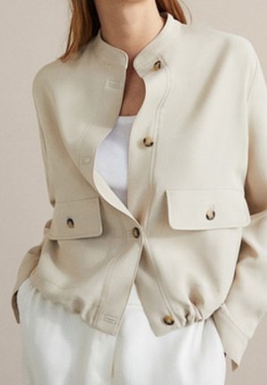 Vrouw draagt een beige blousejas met klepzakken aan de voorkant over een wit shirt en witte broek, handen in de zakken.