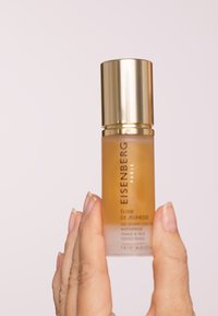 Bouteille Elixir EISENBERG Paris, en verre clair avec un capuchon doré, remplie d'un liquide ambré, étiquetée en texte noir, tenue entre des doigts.
