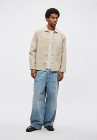 Beżowa bawełniana kurtka z czterema kieszeniami i żółtymi guzikami, w zestawieniu z jasnoniebieskimi szerokimi spodniami i ciemnobrązowymi butami. Gładka faktura.