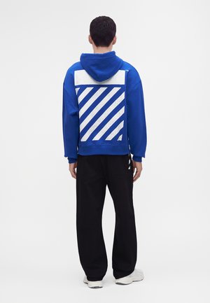 Homme debout de dos portant un sweat à capuche bleu avec des rayures diagonales blanches et un pantalon noir sur un fond uni.