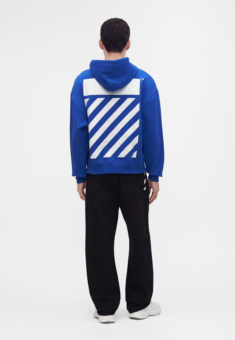 Homme debout de dos portant un sweat à capuche bleu avec des rayures diagonales blanches et un pantalon noir sur un fond uni.