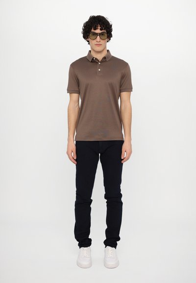 Brown polo shirt with short sleeves, paired with dark pants and white sneakers. Model wears large round sunglasses, standing on a white background.