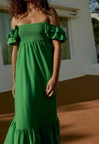 Robe verte décolettée avec des manches volantées, un corsage froncé et un ourlet en étages. Fabriquée en tissu léger, elle présente une texture lisse.