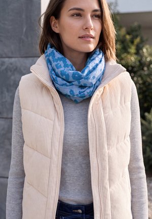 Femme en extérieur portant un gilet matelassé crème, un pull gris et une écharpe bleue à motifs.