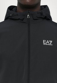 Svart huva jacka med dragkedja fram och dragsko i midjan. Logotypen "EA7 Emporio Armani" visas tydligt i vitt på vänster bröst.