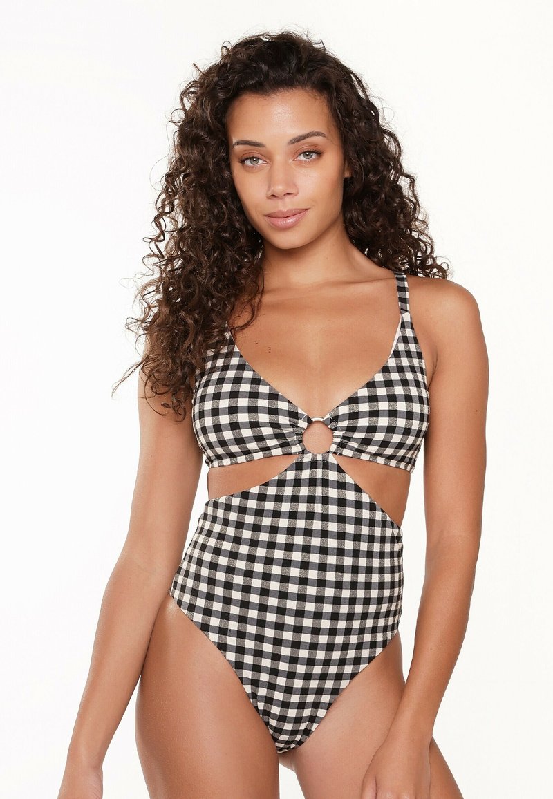 Costume da bagno intero bianco e nero a scacchi, con scollatura profonda a V, fianchi intagliati e un accento circolare al centro. Tessuto morbido al tatto.