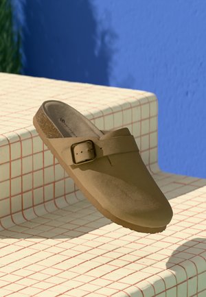 Tan suede slip-on clog with a gold buckle, cork sole, resting on a beige tiled surface with red grid lines, blue wall background.